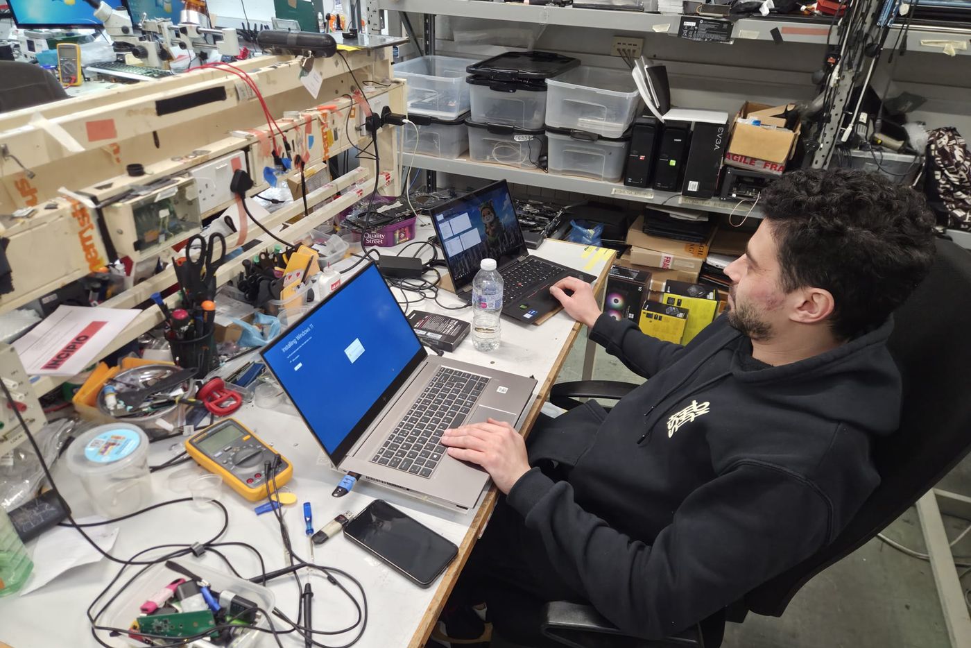 A technician at a workshop bench with two laptops running and a multimeter beside them