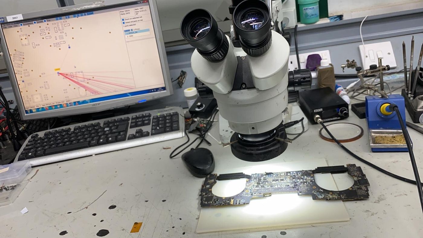 A stereo microscope over a laptop logic board, with a soldering station beside it